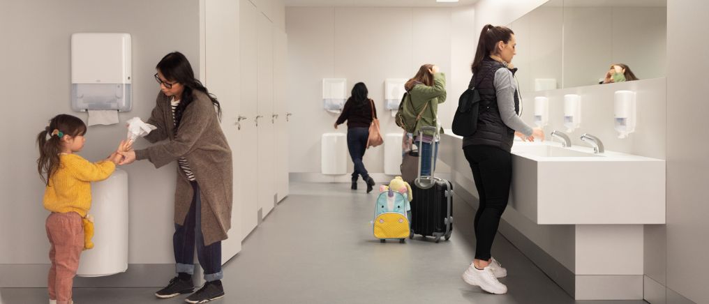 Flera personer använder handfat och pappersdispenser i ett offentligt badrum, inklusive ett barn som får hjälp att torka händerna.