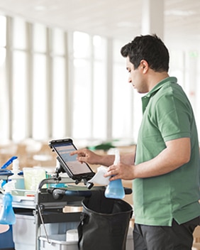 En renholder bruker et nettbrett på en rengjøringsvogn og holder en sprayflaske i hånden i et lyst og moderne rom.