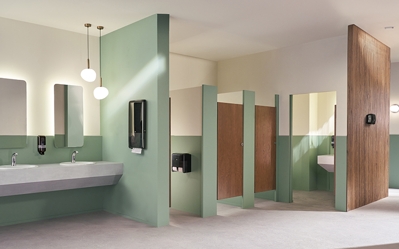 Modern public restroom with light green walls and wooden stall doors, featuring two sinks with mirrors, and soap dispensers. A paper towel and airfreshener dispensers are mounted on the wall.