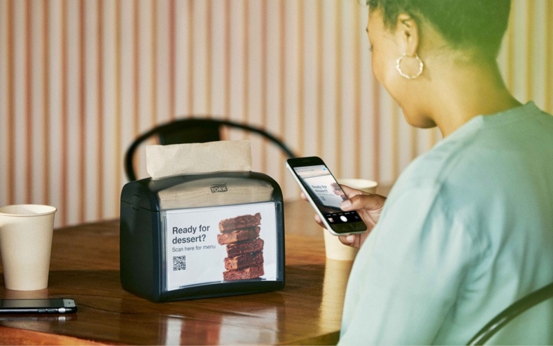 A person seated at a table uses their phone to read a QR-code that is placed on a napkin dispenser