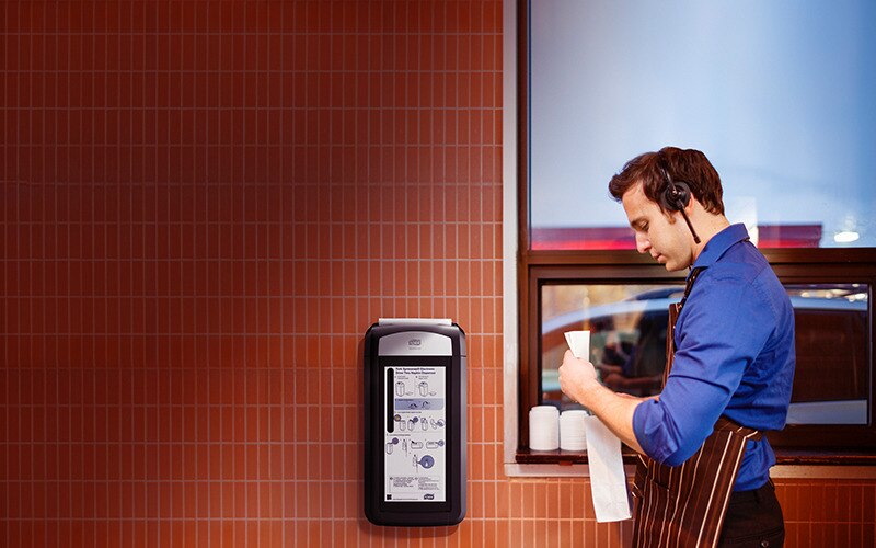 A man in facemask and apron preparing an order in front of a window, there is a Tork dispenser on the wall