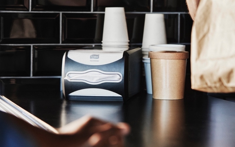 Close up of a Tork napkin dispenser and take-away paper cups on a counter 