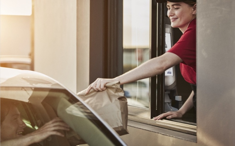 A person working at a drive thru is passing a paper bag out of the window to a person seated in a car 
