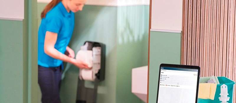 Närbild på en surfplatta på en städvagn och en städare som fyller på en dispenser med toalettpapper