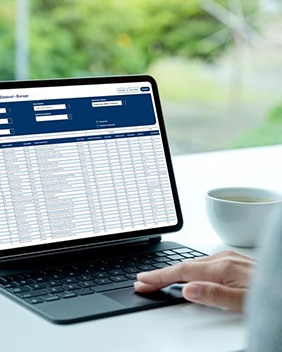 Une personne utilise une tablette avec un tableau de données ouvert à l’écran ; une tasse est posée sur le bureau et de la verdure est visible à l’extérieur.