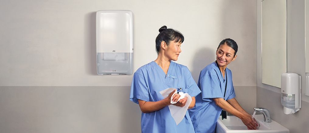 Two healthcare workers in blue scrubs washing and drying hands at a sink in a clinical washroom, with wall-mounted soap and paper towel dispensers. 