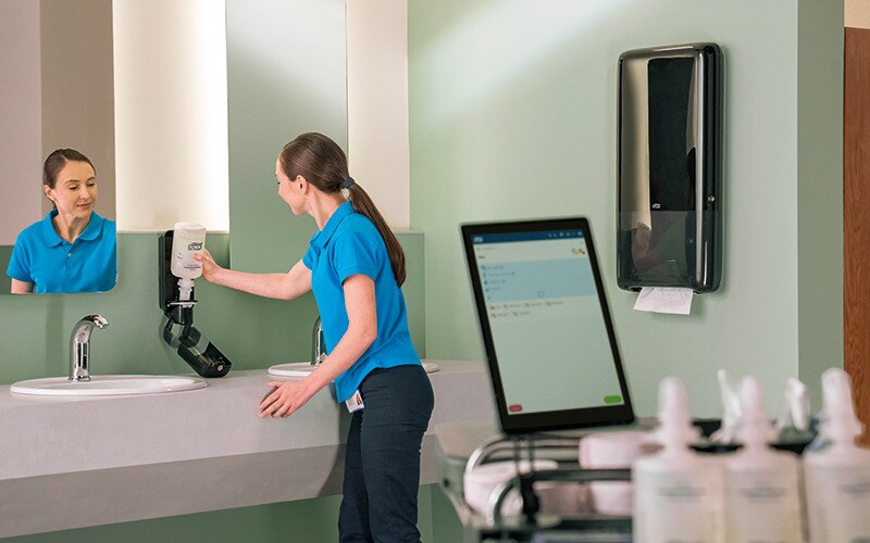 A close-up of a tablet showing workplace hygiene cleaning tasks as a cleaner refills a soap dispenser in the background.​