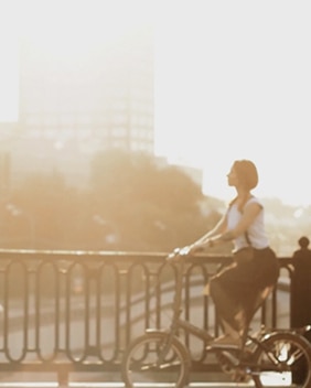 A person riding a bicycle along a bridge railing in warm, bright sunlight, with a softly blurred cityscape in the background.