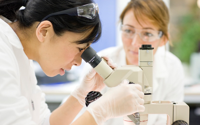 Eine Frau blickt in ein Mikroskop und eine andere Frau steht im Hintergrund