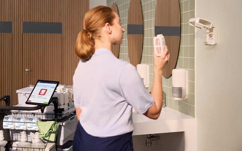Rengøringsassistent står på et toilet ved en rengøringsvogn med rengøringsprodukter med en airfreshener refill i hånden