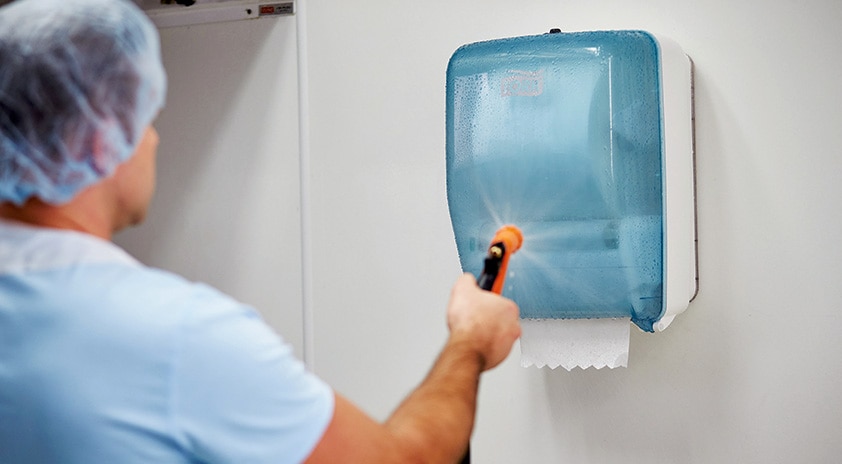 A man wearing a hair net hosing down a Tork Washstation dispenser in a food processing workplace.
