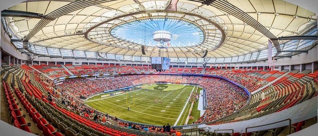 Wide panoramic view of a large indoor stadium filled with spectators, overlooking a football field under a bright, domed roof.