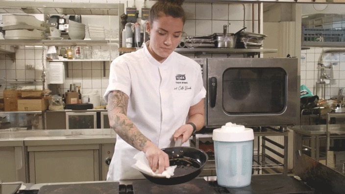 A chef in a kitchen wipes a frying pan with a paper towel, standing beside a large container of cleaning wipes. 