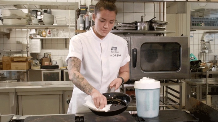 A chef in a kitchen wipes a frying pan with a paper towel, standing beside a large container of cleaning wipes. 