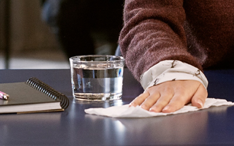 a hand with a paper hand towel wiping a desk, a glass of water, a note book and a pen.