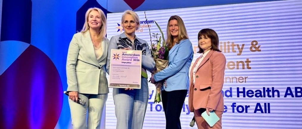 Four people standing on a stage as one person holds an “Amsterdam Innovation Award 2026” certificate and a bouquet of flowers. 