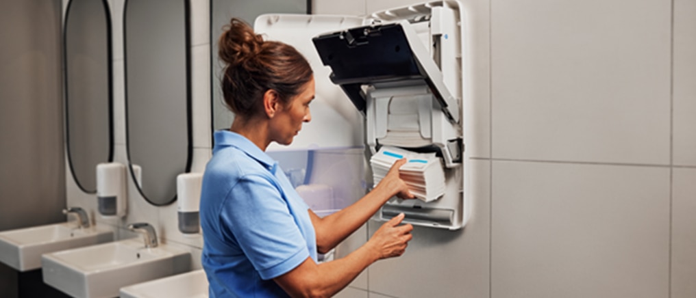 A cleaning person refills a wall-mounted dispenser with paper towels in a public restroom. 