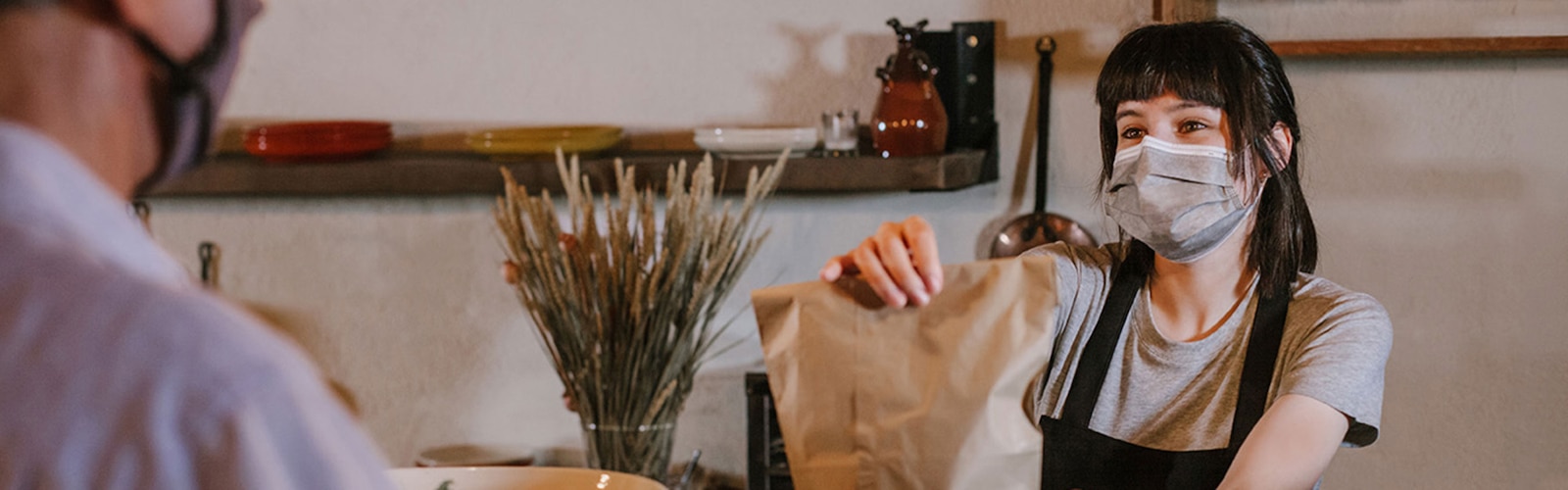 Kvinde med ansigtsmaske, som giver en pose med take-away mad til en kunde