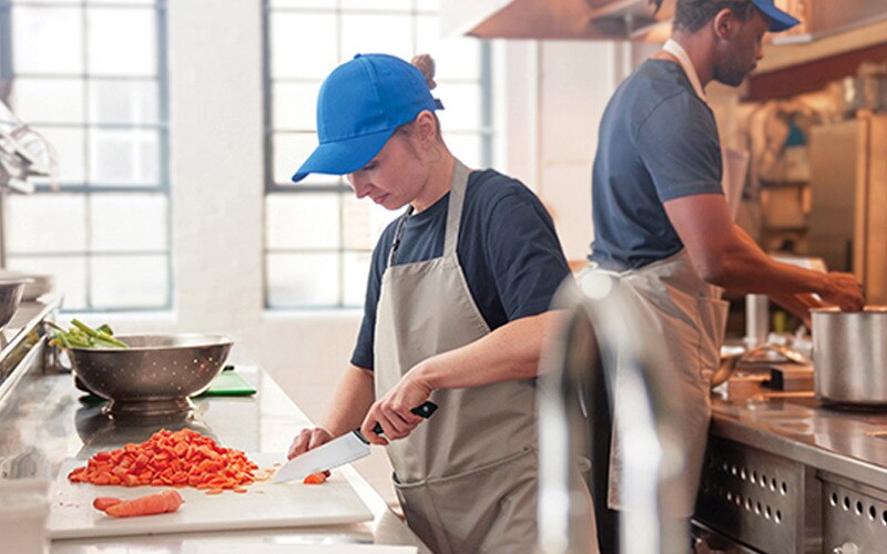  en person med blå keps och förkläde skär grönsaker i ett restaurangkök