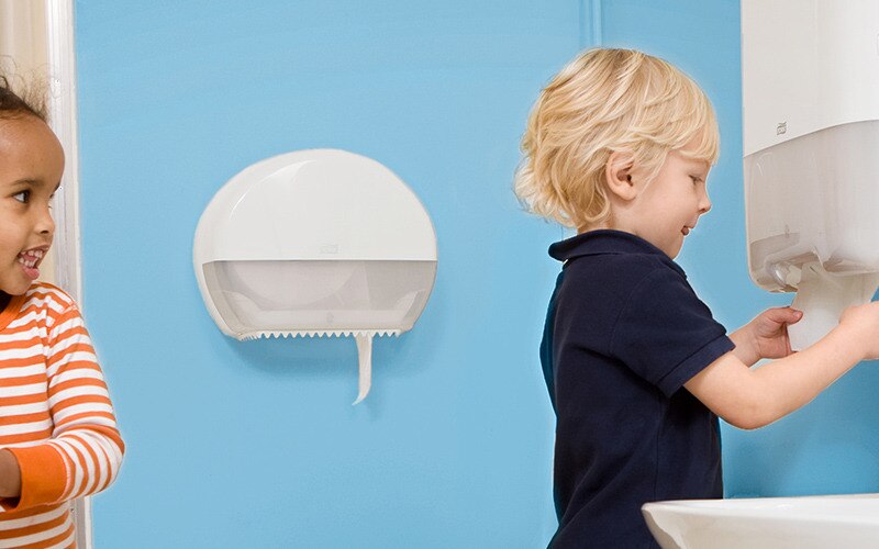 Twee kinderen in een sanitaire ruimte; één veegt haar handen af terwijl een jongen een papieren handdoek uit een dispenser pakt