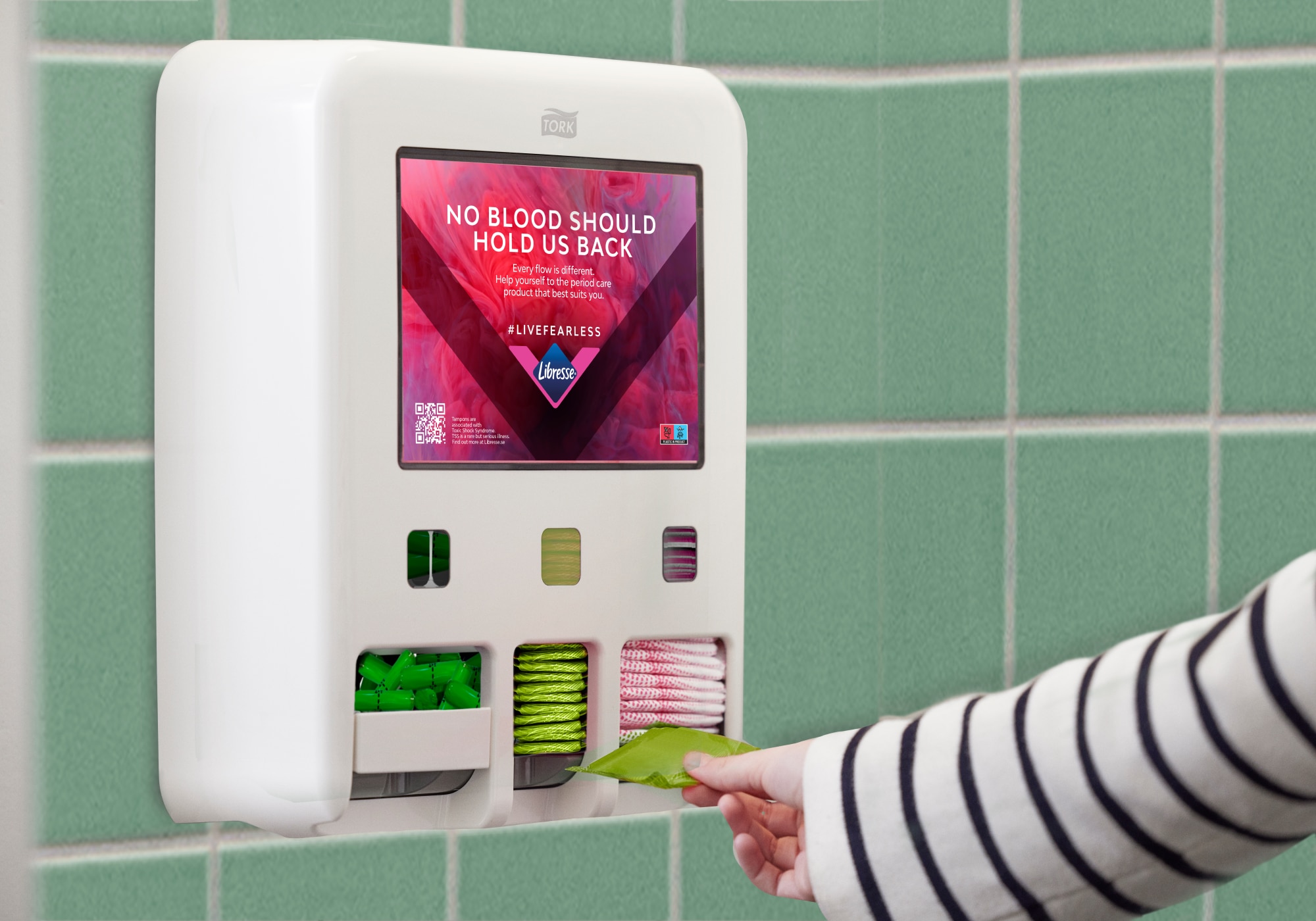 Osoba si odebírá menstruační potřebu z Tork zásobníku na vložky a tampony instalovaného na obložené stěně. Na displeji je vidět zpráva „Menstruace by nás neměla nijak omezovat“.