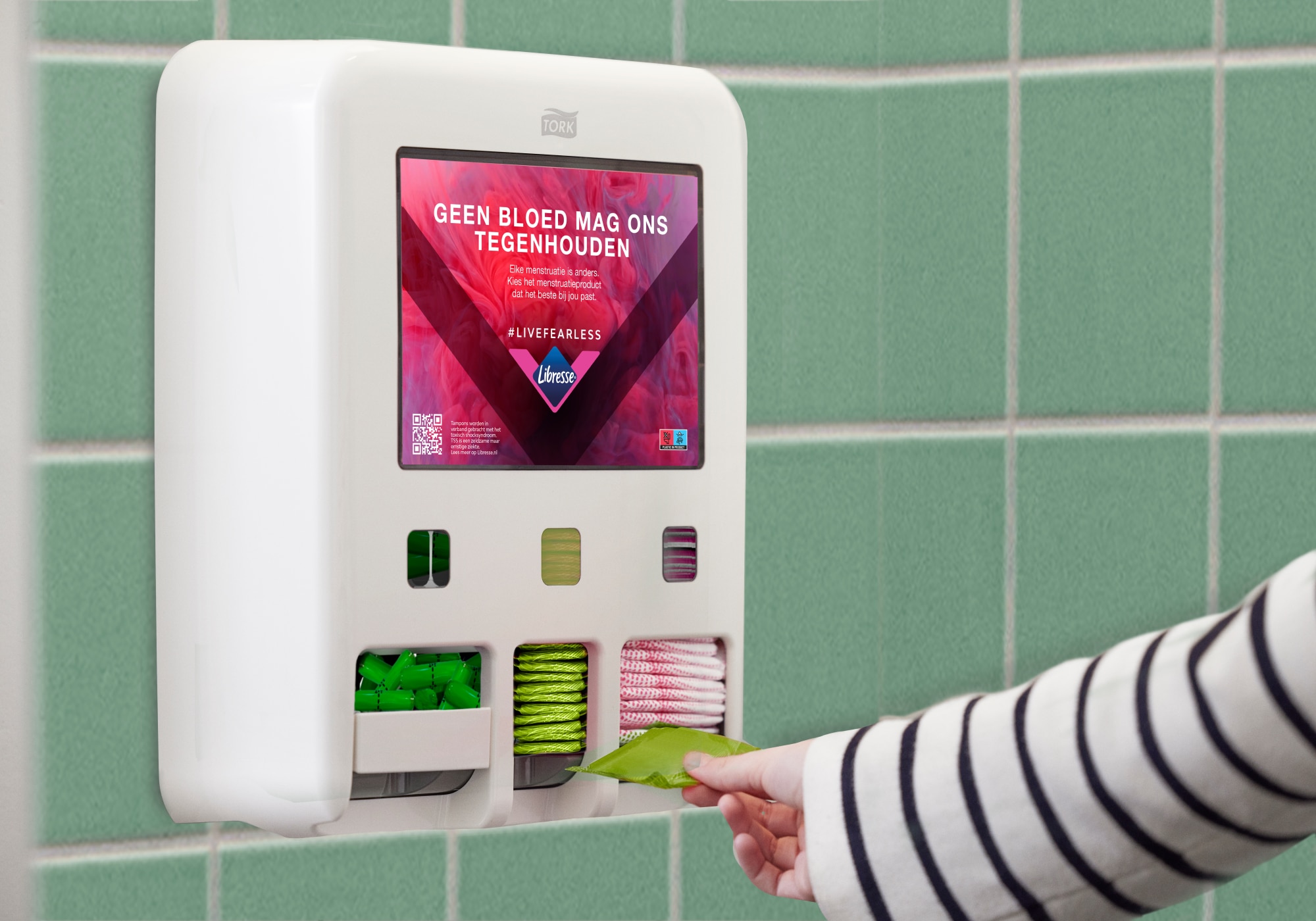Een persoon die een menstruatieproduct pakt uit een Tork Dispenser voor Menstruatieproducten die op een betegelde muur is gemonteerd, met een scherm waarop de boodschap wordt weergegeven "Niets houdt ons tegen, ook menstruatie niet". Een persoon die een menstruatieproduct pakt uit een Tork Dispenser voor Menstruatieproducten die op een betegelde muur is gemonteerd, met een scherm waarop de boodschap wordt weergegeven "Niets houdt ons tegen, ook menstruatie niet".