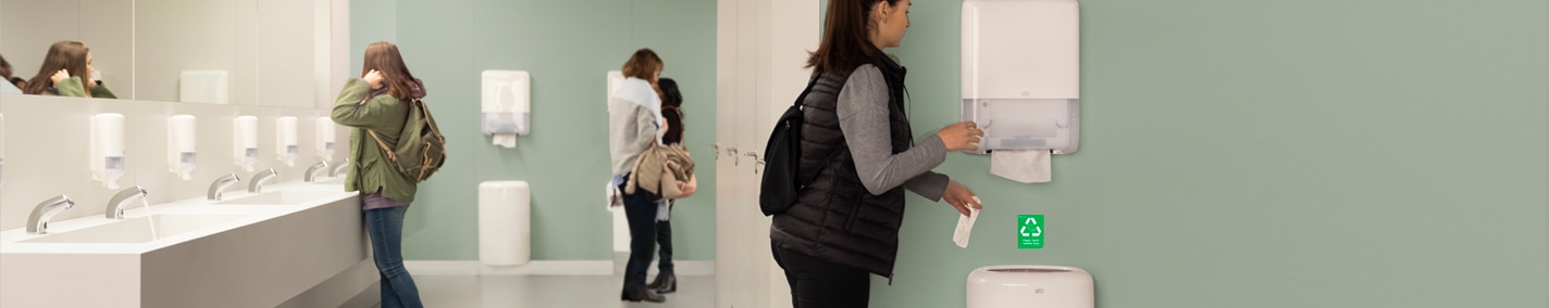 Offentligt badrum med flera kvinnor. Till vänster finns en rad handfat med speglar ovanför. I mitten står en kvinna vid en pappershanddukshållare och tar ut en pappershandduk. I bakgrunden syns två personer, varav den ena håller ett barn.