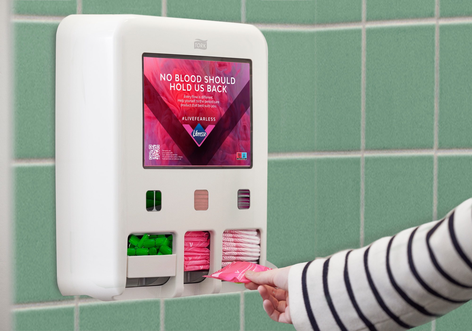 En person tar et mensprodukt fra en Tork Period Care Dispenser montert på en vegg med fliser. På dispenseren står det «No blood should hold us back».