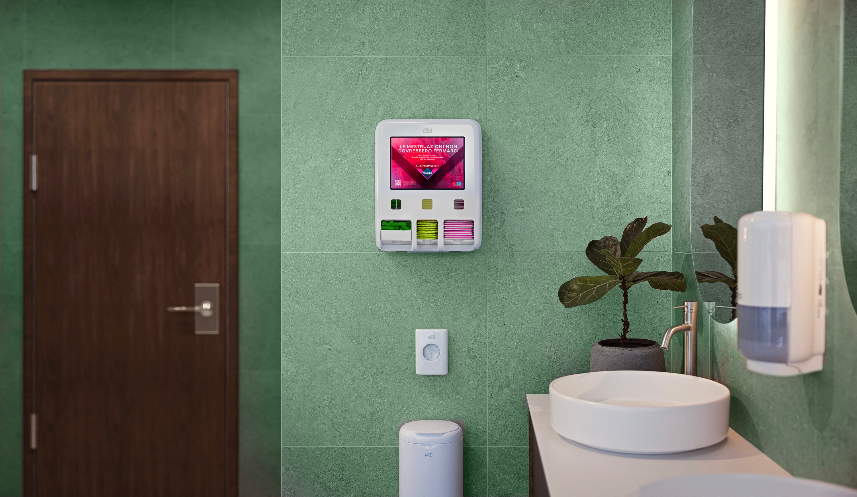 Dispenser Tork di prodotti per l’igiene mestruale montato su una parete con piastrelle verdi dell’area bagno, con un messaggio visibile e diversi vani di stoccaggio, accanto a un lavabo, una pianta e a un cestino. Dispenser Tork di prodotti per l’igiene mestruale montato su una parete con piastrelle verdi dell’area bagno, con un messaggio visibile e diversi vani di stoccaggio, accanto a un lavabo, una pianta e a un cestino.