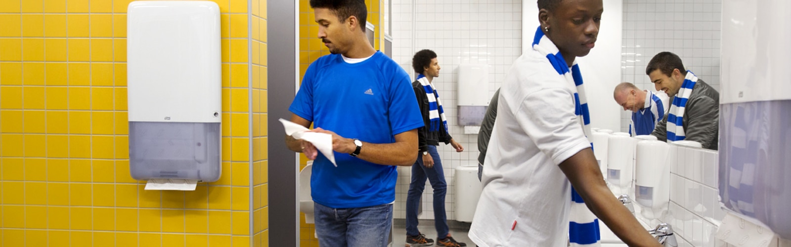 En gruppe menn vasker og tørker hendene i et toalettrom