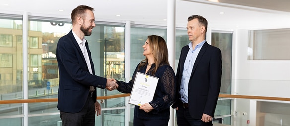 Drei Personen stehen in einem modernen Gebäude; zwei von ihnen schütteln sich die Hand, während eine Person eine Urkunde hält.