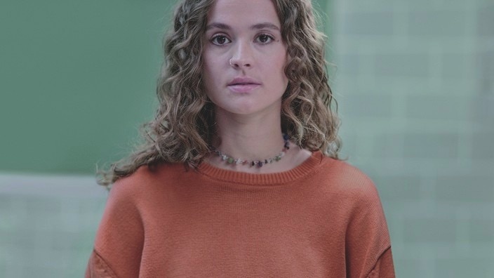 A person with eczema wearing an orange jumper and a beaded necklace, standing in front of a green tiled background. A person with eczema wearing an orange jumper and a beaded necklace, standing in front of a green tiled background.