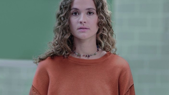 A person with eczema wearing an orange jumper and a beaded necklace, standing in front of a green tiled background. A person with eczema wearing an orange jumper and a beaded necklace, standing in front of a green tiled background.
