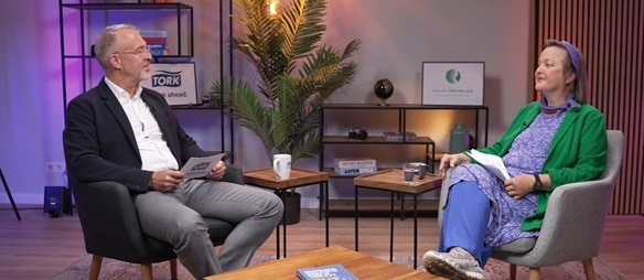 Zwei Personen sitzen in einem Studio und führen ein Gespräch; sie halten Unterlagen in der Hand und sind von Möbeln, Beleuchtung und Dekoration umgeben.