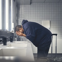A man in a dark hoodie bending over a sink in a tiled bathroom, washing his face with water. A man in a dark hoodie bending over a sink in a tiled bathroom, washing his face with water.