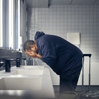 A man in a dark hoodie bending over a sink in a tiled bathroom, washing his face with water. A man in a dark hoodie bending over a sink in a tiled bathroom, washing his face with water.