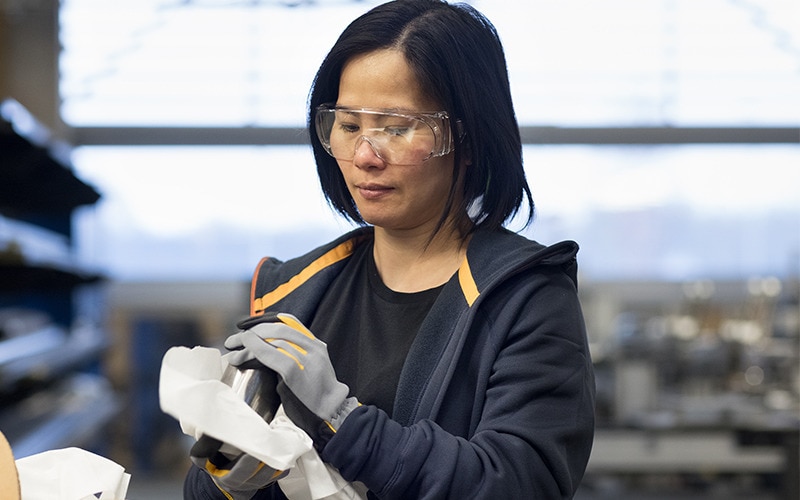 A female industry worker wearing safety glasses is wiping a piece of metal with a paper towel.
