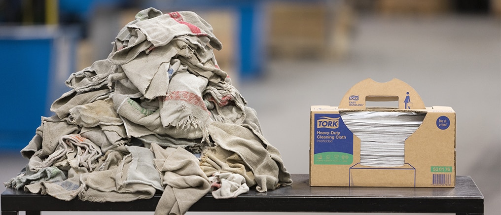 On a table, a pile with textile rags to the left compared to a box Tork Heavy-Duty Clenaing Cloths to the rigt On a table, a pile with textile rags to the left compared to a box Tork Heavy-Duty Clenaing Cloths to the rigt