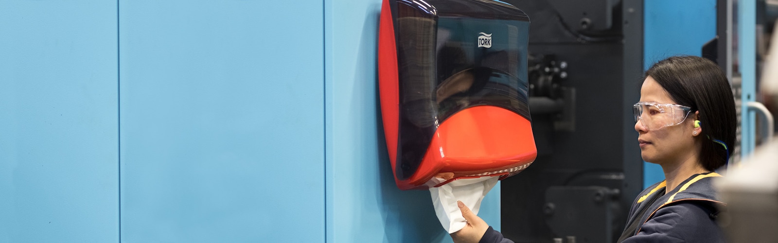 A female industry worker wearing safety glasses is pulling a paper towel out of a Tork Performance dispenser.