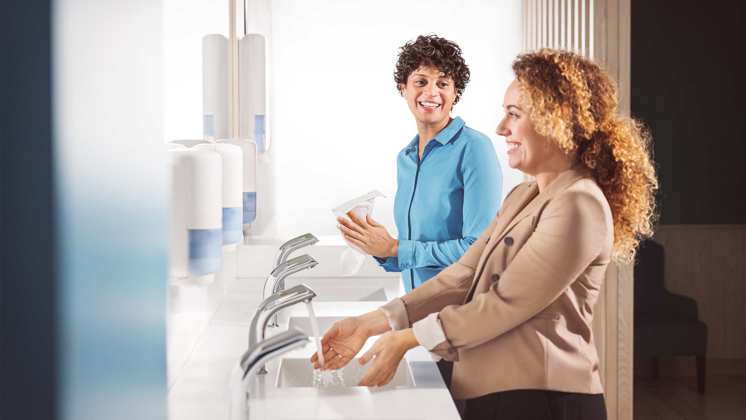 Deux personnes utilisant des distributeurs sans contact de savon et d’essuie-mains le long d’une rangée de lavabos modernes dans une salle de bain bien éclairée.