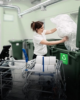 Un membre du personnel de nettoyage recycle des essuie-mains en papier usagés en vidant un sac dans le conteneur d’un local de recyclage.
