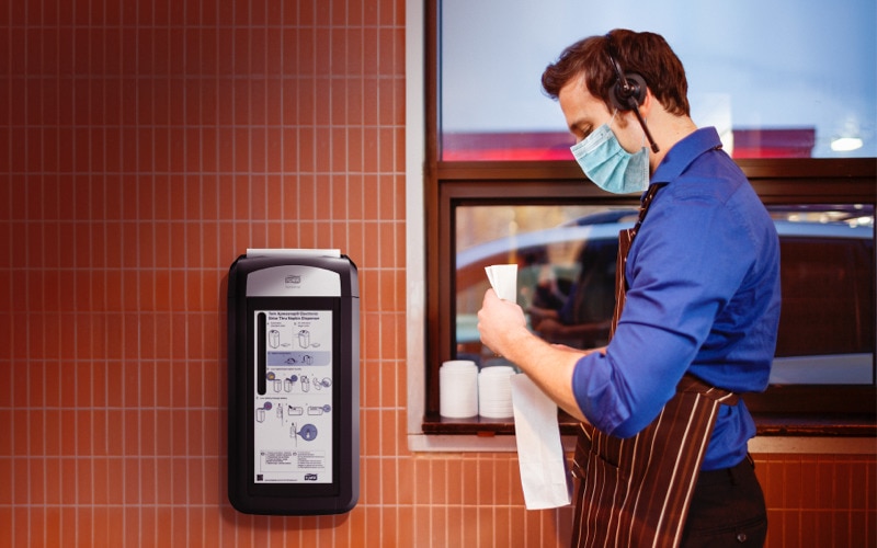 A man in facemask and apron preparing an order in front of a window, there is a Tork dispenser on the wall