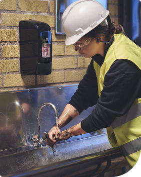 Ein Industriearbeiter wäscht sich die Hände unter einem Wasserhahn, hinter ihm befindet sich ein Spender mit dem Tork System für Handreiniger und Handcreme. Ein Industriearbeiter wäscht sich die Hände unter einem Wasserhahn, hinter ihm befindet sich ein Spender mit dem Tork System für Handreiniger und Handcreme.