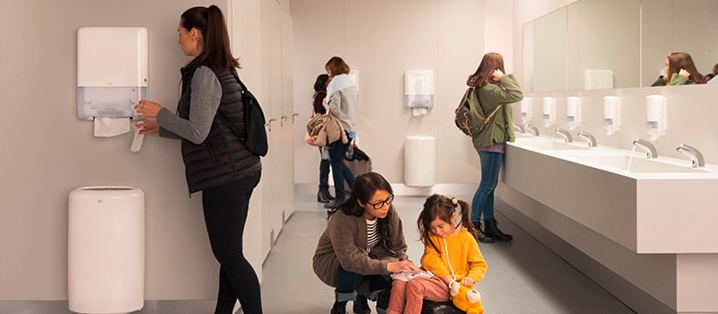 Modern public restroom with people using soap and hand towel dispensers, sinks, and walking toward stalls. Modern public restroom with people using soap and hand towel dispensers, sinks, and walking toward stalls.