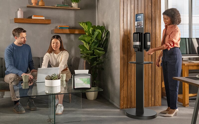 Two employees seated in an office breakroom area looking at a laptop. Next to them another employee is standing next to a hygiene stand.