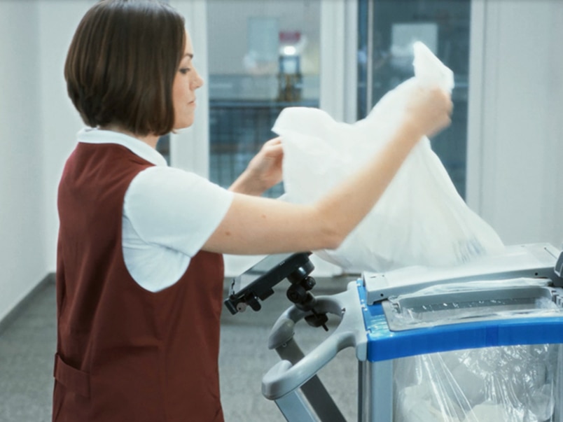 A female cleaner throwing a waste bag into a bag on a cleaner cart