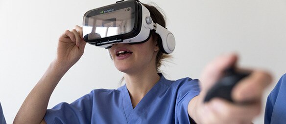 Eine medizinische Fachkraft in blauer Arbeitskleidung verwendet ein Virtual-Reality-Headset und hält einen Controller in der Hand.