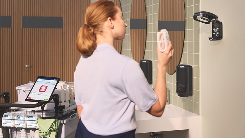 Cleaner standing next to a cleaning trolley with cleaning supplies in a restroom, holding an air freshener refill