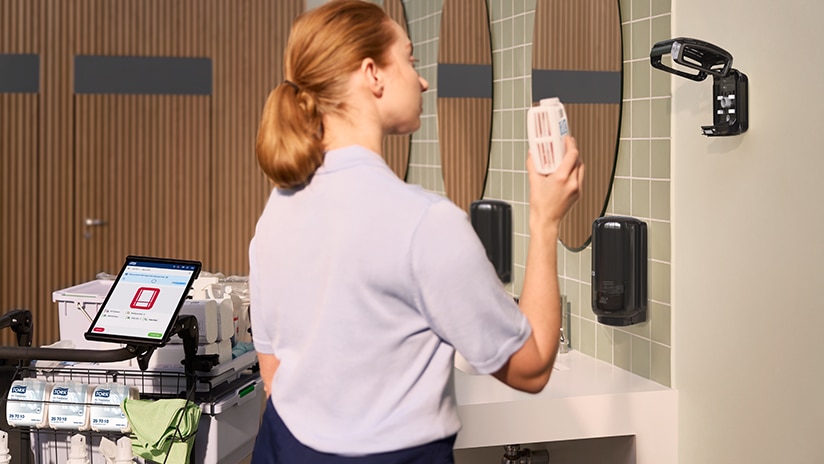 Cleaner standing next to a cleaning trolley with cleaning supplies in a restroom, holding an air freshener refill Cleaner standing next to a cleaning trolley with cleaning supplies in a restroom, holding an air freshener refill