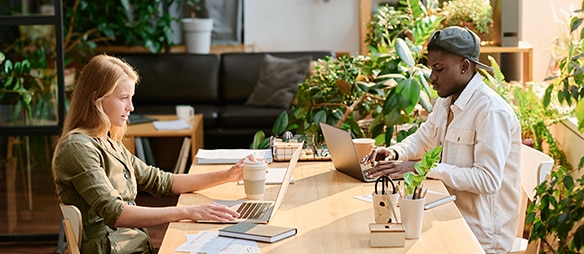 Une femme et un homme assis à une table travaillant sur leur ordinateur portable avec une tasse de café chacun; en arrière-plan, un canapé noir et des plantes vertes